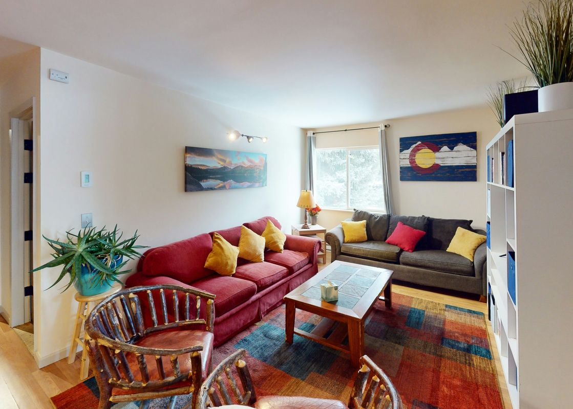 The living room of Beaver Creek Basecamp features a red couch with yellow pillows, a brown couch with yellow and red pillows, a coffee table with a book, and a rug with a blue and brown pattern.
