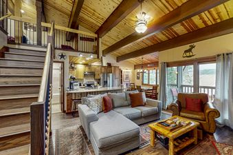 A spacious living area with vaulted wooden ceilings, a gray sectional sofa, and a wooden coffee table, featuring a kitchen in the background and a staircase leading to an upper loft.