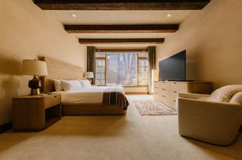 A cozy bedroom with a large bed, wooden furniture, and a window looking out onto a building and trees.