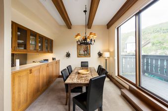 The dining room at Plaza Lodge 3 Bedroom Gold Unit 4 features a rustic wooden table with black leather chairs, illuminated by a chandelier, with a view of a mountainous landscape from a large window.