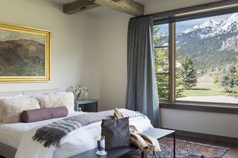 The bedroom of Fish Creek Lodge 63 features a bed with white linens and furry pillows, a nightstand, and a large window overlooking snow-capped mountains and pine trees.