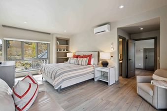 A spacious bedroom at Village Hall 09 features a comfortable bed with a striped comforter and numerous pillows, a sleek white nightstand, a built-in bookshelf, and a large window offering scenic outdoor views.