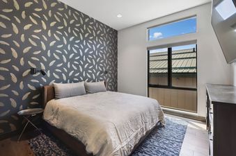 A modern bedroom with a feather-patterned accent wall, a large bed with gray bedding, a metallic side table, and a window offering an exterior view.