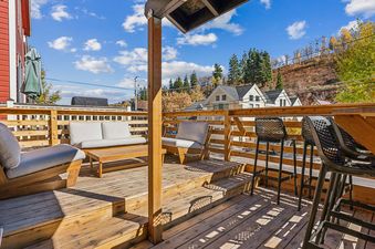 A spacious wooden outdoor deck at Daly Cottage featuring lounge seating, a coffee table, and a bar counter with stools, all overlooking autumn hills under a blue sky.