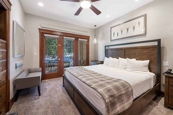 A primary bedroom in The Charter G1305 featuring a king-sized bed, a gray bench, leaded glass doors, and a framed art piece of deer.