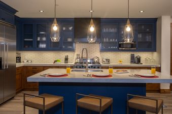 A modern kitchen with navy blue cabinetry, stainless steel appliances, a large island with breakfast settings, and elegant pendant lighting.