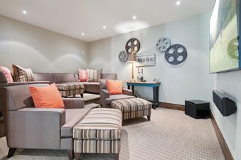 A cozy home cinema room at Winter Watch Estate featuring tiered sectional seating with orange and striped pillows, film reel wall decor, a large flat-screen TV with a sound system, and soft lighting.
