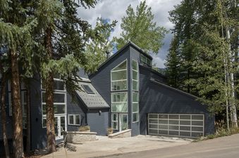 A modern, dark grey house with large windows and a multi-level design is nestled among tall trees, featuring a garage and a sloped driveway.