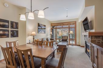 The dining area of Capitol Peak Lodge 3207 features a wooden table with chairs, a view of the mountains from a large window, and a fireplace in the background.