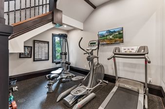 The home gym at Sky Ridge Retreat, featuring an elliptical, treadmill, and weight machine, with a TV mounted on the wall and a window offering a green outdoor view.