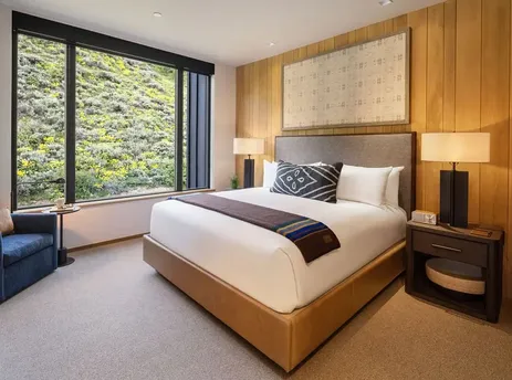 A king guest room at The Sylvan Lodge features a king-sized bed with white linens, a comfortable armchair by a large window overlooking a lush green hillside, and warm wood-paneled walls.