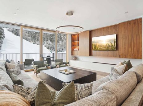 A modern living room with a large sectional sofa, two olive green armchairs, and a dark wooden coffee table, featuring floor-to-ceiling windows overlooking a snow-covered mountain slope with pine trees.