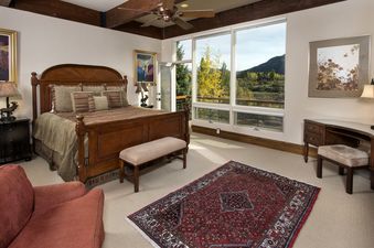 A spacious bedroom at 185 Nighthawk Dr with a large wooden bed, two bedside tables with lamps, a bench, a red armchair, a desk, and large windows offering a scenic view of fall foliage and mountains.