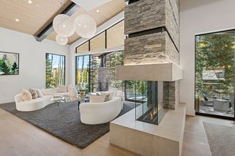 A modern living room at All In at The Colony with a stone and glass fireplace, cream sofas, and large windows looking out onto a forest and mountains.
