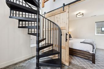 A black spiral staircase ascends alongside a natural wood barn door, revealing a bedroom with a bed and window in Quarry A4.