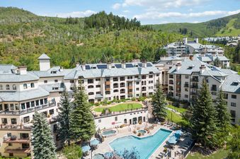An aerial view of The Charter C1105, a multi-story building surrounded by green mountains and evergreen trees, with an outdoor pool and hot tubs.