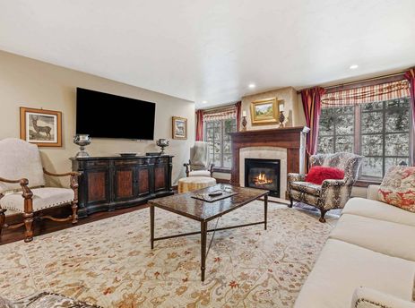 A luxurious living room with a large TV, a fireplace with a mantel, and two windows showcasing a snowy outdoor scene.