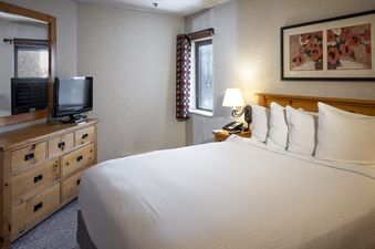 A cozy bedroom in the Building 4 Two Bedroom Breckenridge Suite with a bed, dresser with TV and mirror, a nightstand with a lamp, and a window with a patterned curtain.