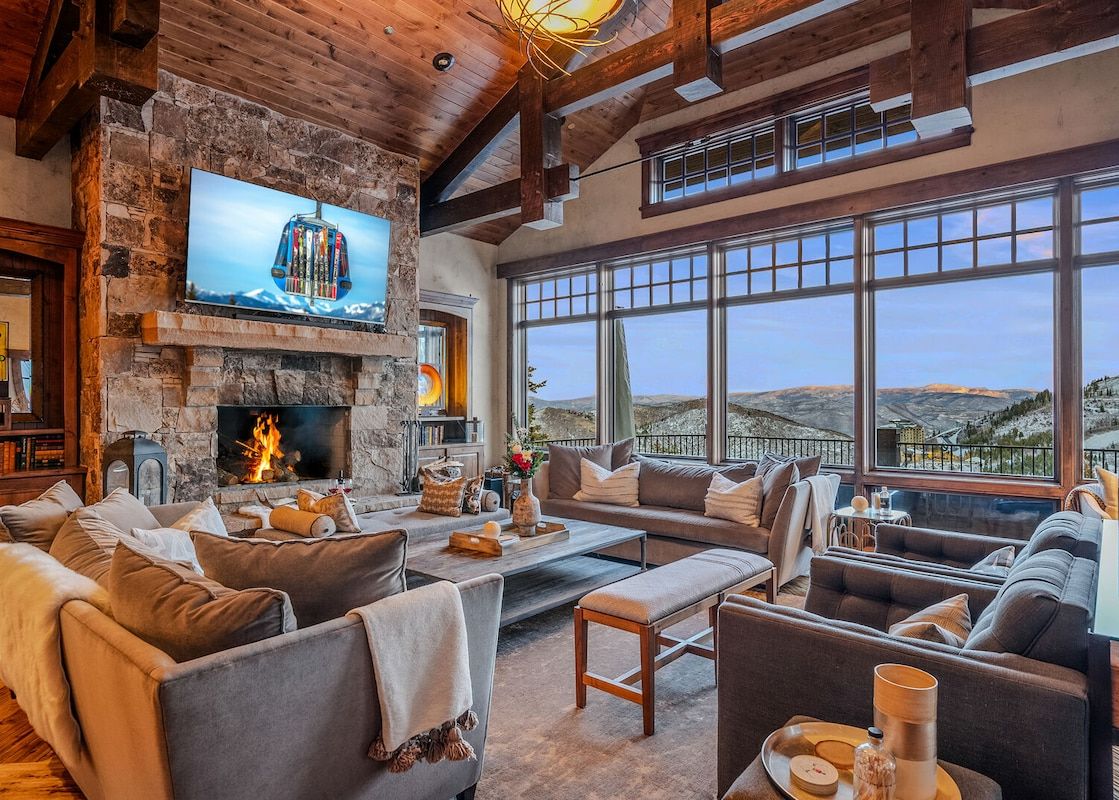 Warmly lit living room with stone fireplace, comfortable grey seating, and a large window looking out onto mountain views.
