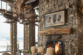 The grand dining area at Phillips Ridge features a large wooden table with seating for eight, an antler chandelier, and a stone fireplace with a painting above, all set against a backdrop of snowy outdoor views.