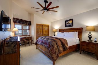 A well-appointed bedroom at Chateau Chamonix CX233 features a wooden bed with a colorful comforter, a desk by a window, and a wall-mounted TV.