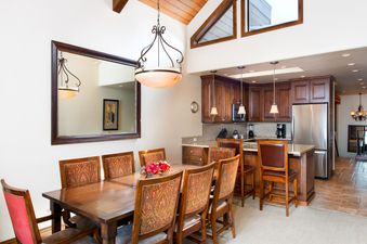 A formal dining room with a long wooden table and chairs with colorful upholstery next to a modern kitchen with dark wood cabinets and stainless steel appliances.