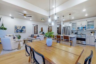 A dining table with chairs, lit by hanging pendant lights, sits in front of a modern kitchen with light grey cabinetry and stainless steel appliances.