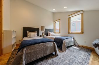 Two twin beds with dark wooden headboards, blue throws, and patterned pillows sit on a blue patterned rug in a light-colored room with two windows.