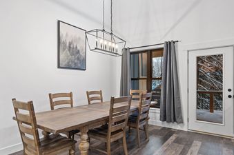 A rustic wooden dining table and chairs are illuminated by a geometric chandelier, with a window and glass-paned door revealing a snowy outdoor scene.