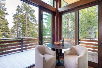 A cozy balcony seating area at West Shore Hideaway with two armchairs, a table with wine and snacks, and a scenic view of snow-covered pine trees through large glass windows.