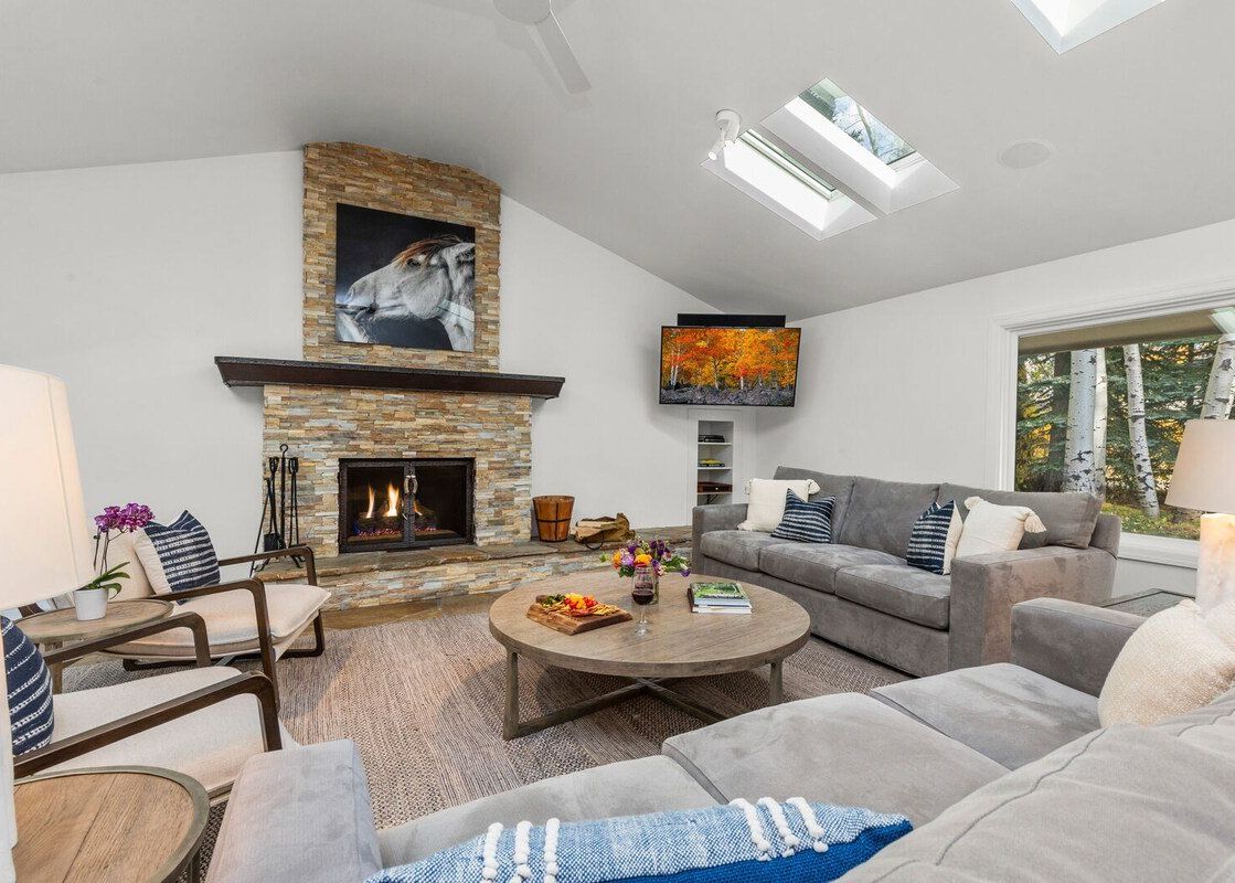 A spacious living room featuring a stone fireplace, gray sofas, skylights, and a window with a forest view.