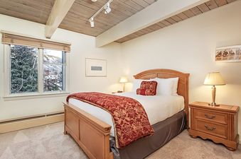 A cozy bedroom at Chateau Chaumont Unit 9 features a queen-sized bed with elegant red and gold bedding, flanked by two nightstands with lamps, and a window offering a scenic winter view.