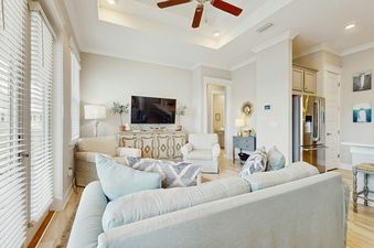 A bright and airy living area featuring light-colored walls, comfortable sofas with decorative pillows, a flat-screen TV on a console, and large windows with white blinds.