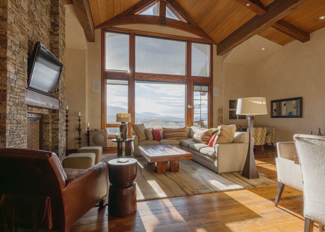 A spacious living room with vaulted ceilings and large windows overlooks a mountain landscape, featuring a stone fireplace, sectional sofa, leather armchair, and wooden furniture.
