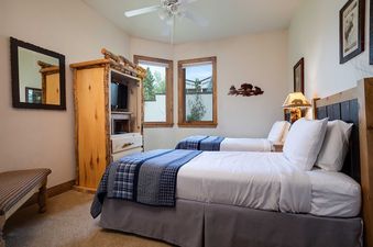 The image shows the second bedroom at Chateau Chamonix CX311, featuring two beds with blue and white plaid bedding, a rustic log-framed armoire with a television, and large windows with an exterior view.