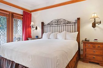 A king-size bed with white linens and a wrought-iron headboard is flanked by wooden nightstands and wall-mounted lamps, with red curtains framing decorative windows.