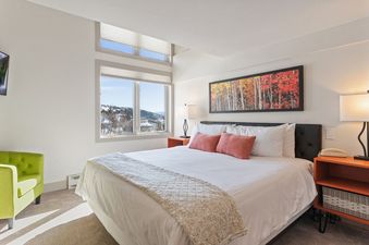 A bedroom with a white bed, two red pillows, and a window with a view of a snowy mountainside