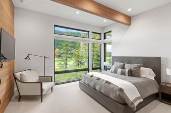 A modern bedroom with a king-sized bed, large windows showcasing a golf course and mountain view, and a cozy armchair with a floor lamp.