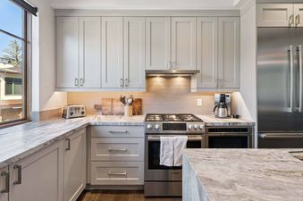 A modern kitchen with light gray cabinets, marble countertops, and stainless steel appliances, including a gas range, microwave, and refrigerator.