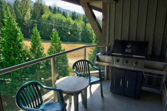 A private balcony at Aspens on Blackcomb Unit 552 features a barbecue grill, a small table, and two chairs, all set against a backdrop of lush green trees and distant mountains under a bright sky.