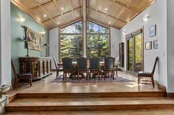 A dining room with a wooden table and chairs, a large window with a view of trees, and a wooden ceiling.