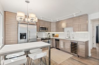 A modern kitchen with light wood-grain cabinets, white countertops, and stainless steel appliances, featuring a high-top dining table with white bar stools.