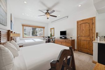 A bright bedroom in The Charter F255 with two queen beds, a ceiling fan, a dresser with a TV, and a small dining table next to a window with an outdoor view.