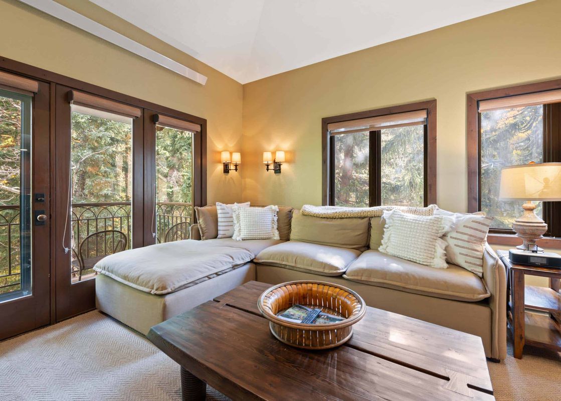 A cozy lounge area in The Charter C2105 featuring an L-shaped sofa, a wooden coffee table, and French doors leading to a balcony with a forest view.