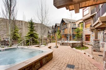 An outdoor patio with hot tubs, a wooden walkway, a stone wall and a red brick paver walkway.  The mountains are in the background.