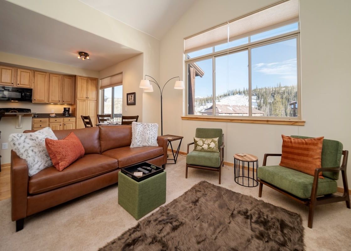 The living area of Trailhead Lodge 731 features a comfortable brown leather sofa, two green armchairs, and a large window overlooking a snowy landscape.