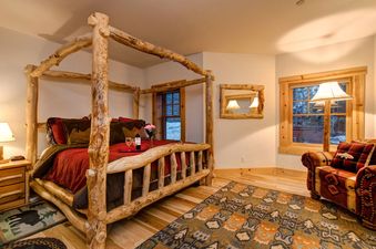 A rustic bedroom at Obsidian Jackson Hole features a large log four-poster bed with red bedding, a cozy armchair, and a window overlooking a snowy landscape.