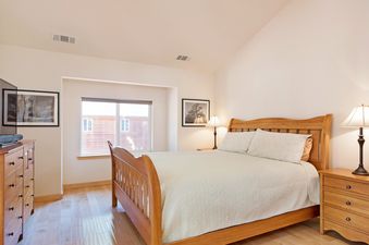 A bright bedroom in North Shore Golden Pine, featuring a wooden sleigh bed with white bedding, two bedside lamps, a window, and a dresser with a TV.