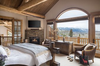 A luxurious bedroom in Mountain View Clear Creek features a cozy bed, a stone fireplace, a flat-screen TV, and large windows with stunning mountain views.
