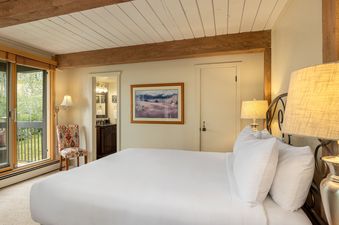 A cozy bedroom with a large bed, exposed wooden beams on the ceiling, a sliding glass door to a balcony, and an en-suite bathroom visible through an open doorway.